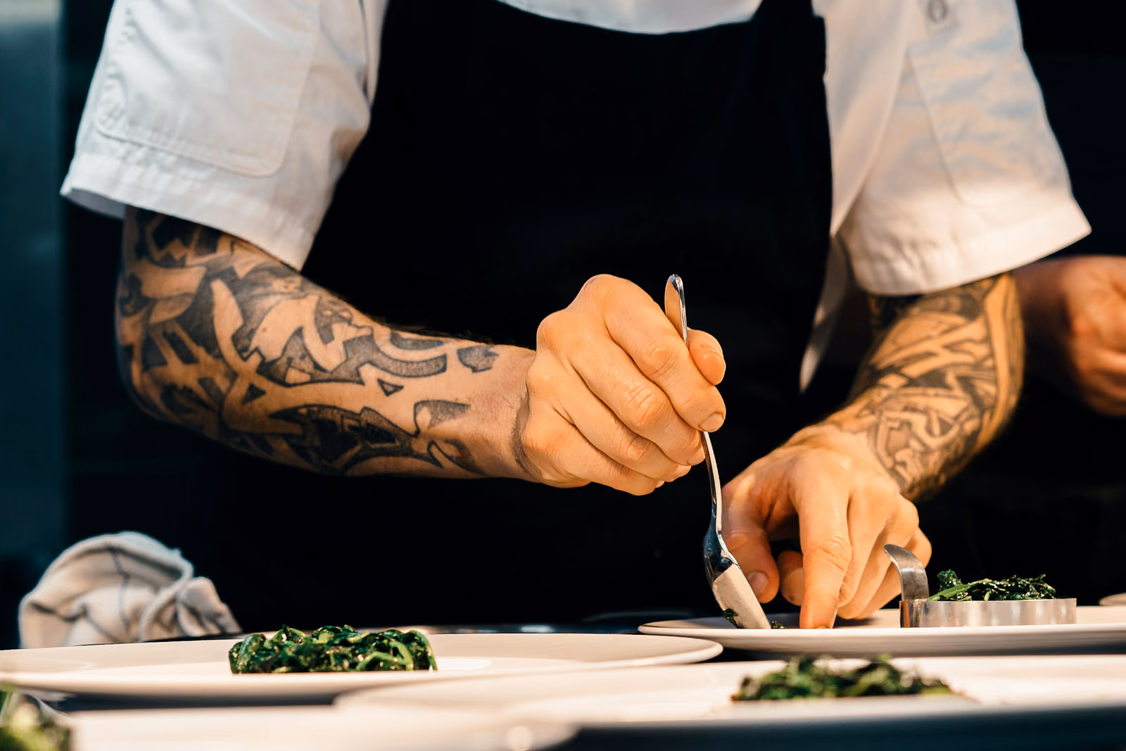 A cook's tattooed forearms and hands plating a dish with tweezers in a dark kitchen